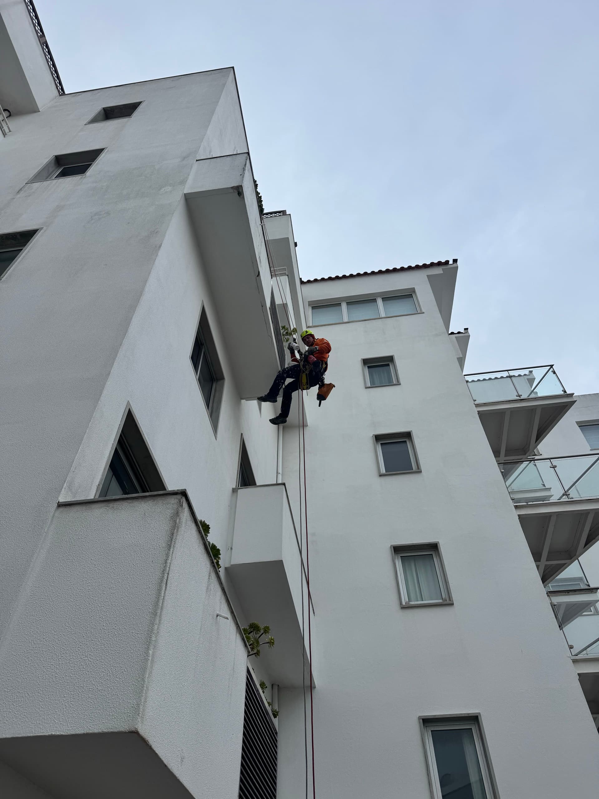Técnico em acesso por cordas em suspensão na fachada de um edifício urbano em altura.